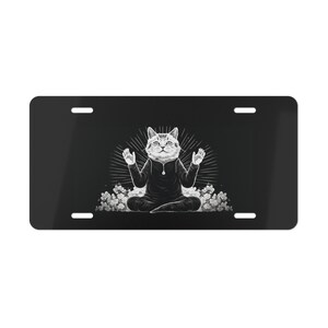 May include: Black license plate with a meditating cat illustration. The cat is in a seated yoga pose, wearing a dark hoodie, with its paws raised. White flowers surround the cat. The background is black.