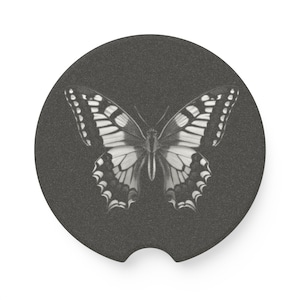 May include: A circular dark gray car coaster featuring a detailed white butterfly illustration centered on the surface. The coaster has a small semi-circular notch at the bottom edge, designed for easy removal from a vehicle cup holder.