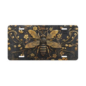 May include: A black license plate with a detailed gold bee design. The bee is centered, with outstretched wings and intricate detailing. The background features floral and scrollwork patterns in gold, creating a luxurious aesthetic.