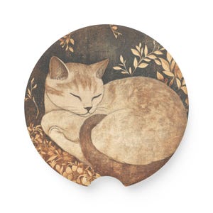 May include: A round car coaster featuring a sleeping cat illustration. The cat is light brown with darker brown stripes and is curled up on a bed of leaves. The background is dark with light brown floral accents.