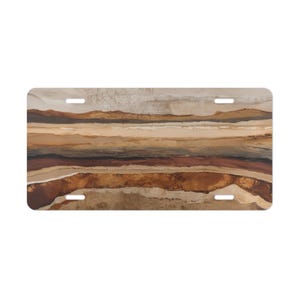 May include: Rectangular license plate featuring an abstract landscape design. The artwork showcases horizontal bands of color in shades of brown, beige, and tan, creating a layered effect. The plate has four pre-drilled holes for mounting.
