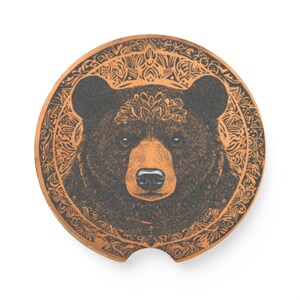 May include: Round car coaster featuring a detailed illustration of a bear's face. The bear is dark brown with a tan muzzle and a decorative crown. The coaster has a brown border with an ornate design.
