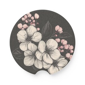 May include: A round car coaster featuring a floral design. The coaster has a dark gray background with a detailed illustration of white flowers and pink buds. The design is a pencil sketch style.