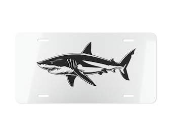 May include: A white license plate featuring a black and white illustration of a shark. The shark is depicted in profile, showcasing its sharp teeth and fins. The plate has four mounting holes.