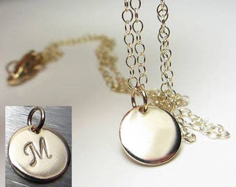 May include: A gold-colored necklace with a delicate chain and a round pendant. The pendant is smooth and shiny, reflecting light. An inset image shows a similar pendant with the letter "M" engraved on it. The necklace is a simple, elegant piece of jewelry.