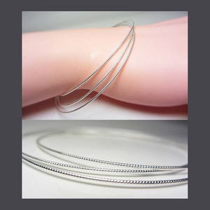 May include: A set of three silver-colored bangles with a textured, beaded design. The bracelets are shown on a light pink surface, with a close-up view of the bracelets below.