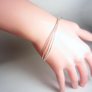 May include: A delicate silver bracelet with three thin bands is worn on a wrist. The bracelet is simple and elegant, reflecting light. The hand is relaxed, showcasing the jewelry's design. The bracelet's minimalist style makes it suitable for everyday wear.