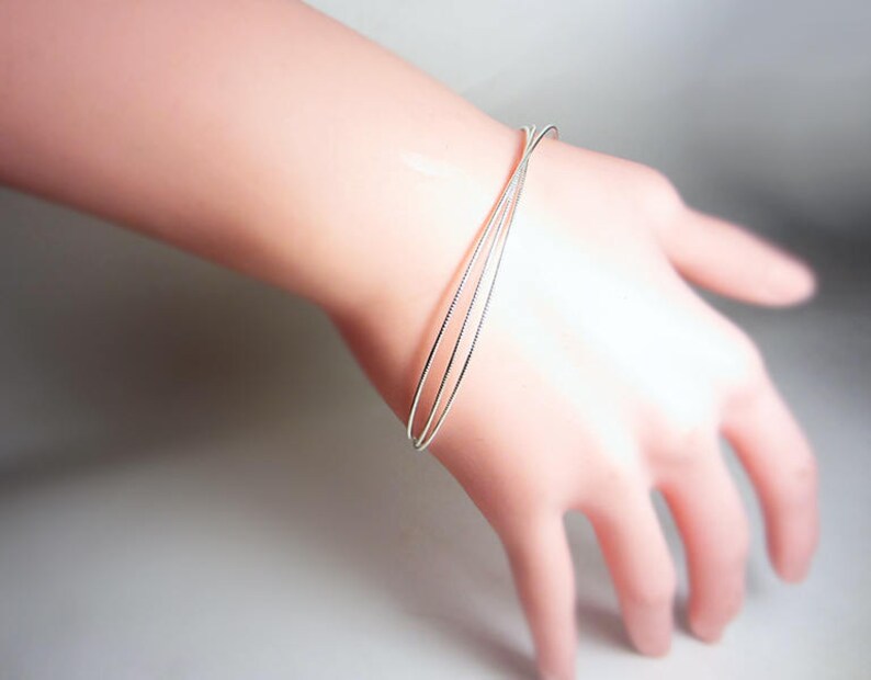 May include: A delicate silver bracelet with three thin bands is worn on a wrist. The bracelet is simple and elegant, reflecting light. The hand is relaxed, showcasing the jewelry's design. The bracelet's minimalist style makes it suitable for everyday wear.