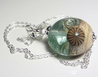 May include: A silver chain necklace with a large, round, glass pendant. The pendant is clear and has a swirl of green, white, and brown inside. The pendant has a textured surface.