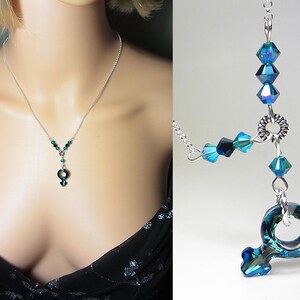 May include: A silver chain necklace with a dark green crystal pendant in the shape of the female symbol. The necklace is adorned with smaller dark green crystals.