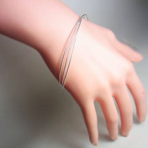 May include: A delicate silver bracelet featuring three thin, parallel bands. The bracelet is worn on a light-skinned hand, showcasing its minimalist design and subtle shine. The bands are approximately 0.1 inches in width.