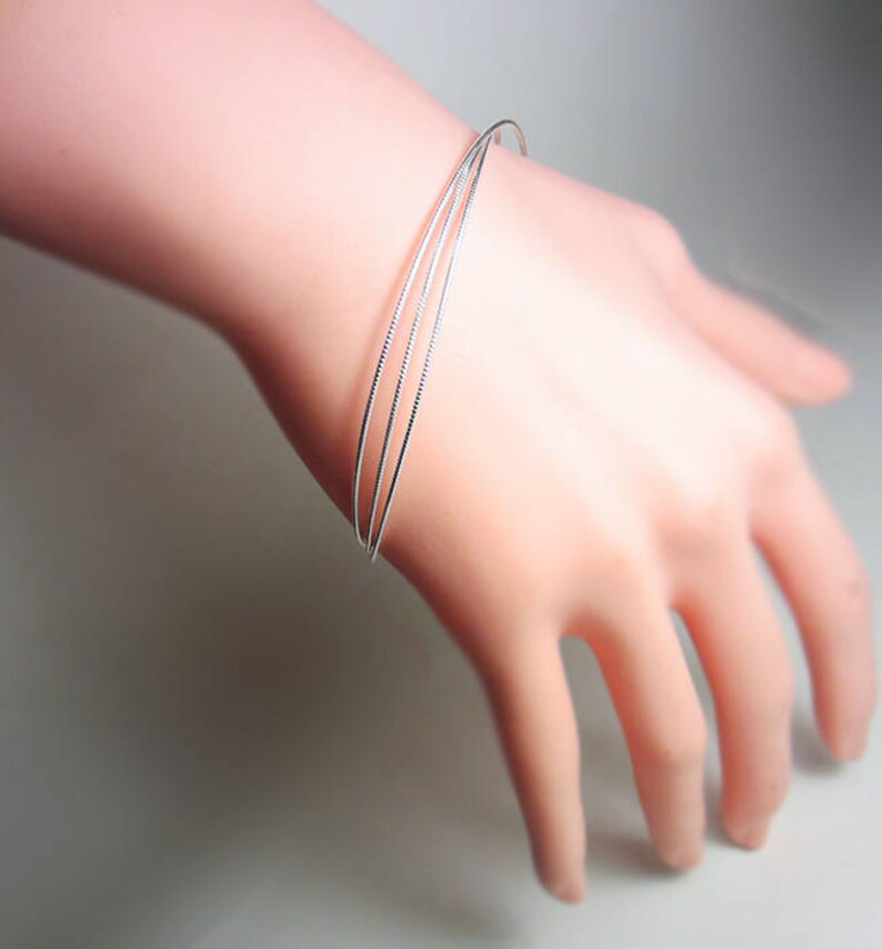 May include: A delicate silver bracelet featuring three thin, parallel bands. The bracelet is worn on a light-skinned hand, showcasing its minimalist design and subtle shine. The bands are approximately 0.1 inches in width.