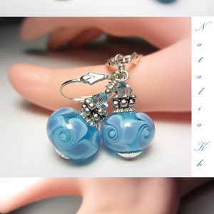 Pu&ograve; includere: Orecchini pendenti con perline di vetro azzurro con un motivo a spirale. Gli orecchini hanno dettagli in metallo argentato e chiusure a leva. Le perline misurano circa 2 cm di diametro.