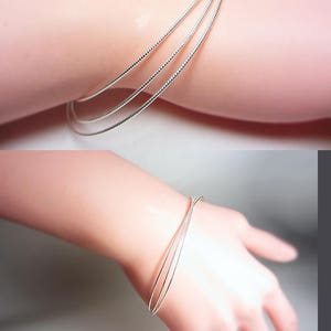 May include: A set of three silver-colored wire bracelets. The bracelets are thin and delicate, with a textured surface. The jewelry is shown on a light-skinned arm.