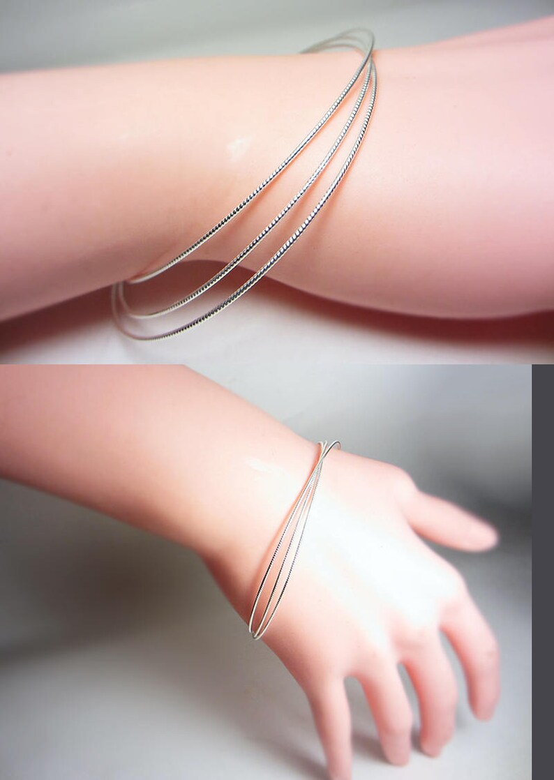 May include: A set of three silver-colored wire bracelets. The bracelets are thin and delicate, with a textured surface. The jewelry is shown on a light-skinned arm.