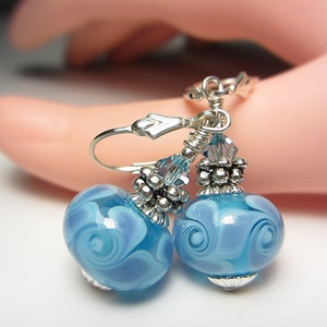 Pu&ograve; includere: Orecchini pendenti con perline di vetro azzurro chiaro e motivi a spirale bianchi. Hanno dettagli in metallo argentato e chiusure a leva. Le perline misurano circa 2 cm di diametro.