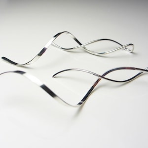 May include: Two silver-toned, abstract earrings with a flowing, wave-like design. The earrings are displayed on a white surface, reflecting the light and creating a sense of movement. The design is minimalist and modern.
