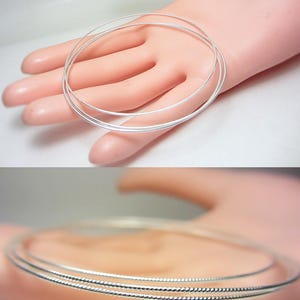 May include: Three silver-colored, thin, wire-like bangles are displayed on a light-toned surface. The bangles are circular and appear to be made of a shiny metal. The image is well-lit, highlighting the reflective quality of the jewelry.