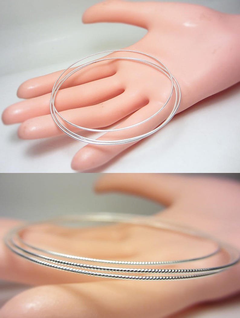 May include: Three silver-colored, thin, wire-like bangles are displayed on a light-toned surface. The bangles are circular and appear to be made of a shiny metal. The image is well-lit, highlighting the reflective quality of the jewelry.