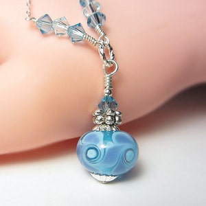 May include: A delicate silver necklace featuring a pendant with a light blue glass bead. The bead has a swirling pattern and is accented with silver and blue crystal beads. The necklace is a beautiful piece of jewelry.