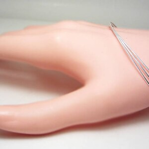 May include: A delicate silver bracelet with three thin, parallel bands. The bracelet is displayed on a light pink mannequin hand, showcasing its minimalist design and elegant simplicity. The bands are approximately 0.08 inches thick.