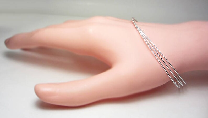 May include: A delicate silver bracelet with three thin, parallel bands. The bracelet is displayed on a light pink mannequin hand, showcasing its minimalist design and elegant simplicity. The bands are approximately 0.08 inches thick.