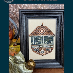 Op de afbeelding: Een ingelijst kruissteekpatroon met een bruin en blauw eikelontwerp met het woord "FALL" in het midden geborduurd. Het kunstwerk staat tegen een crèmekleurige achtergrond en is ingelijst in een zwart houten frame. De afbeelding bevat een gouden eekhoornbeeld.