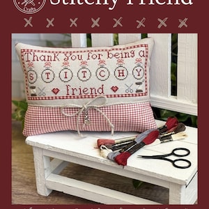 Op de afbeelding: Een kruissteekpatroon getiteld "Stitchy Friend" met een kussen met de tekst "Thank You for being a Friend". Het ontwerp bevat kruissteekelementen, scharen en garenklosjes. Een bundel borduurgaren en een kleine zwarte schaar liggen op een witte bank.