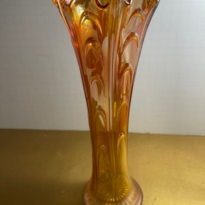 May include: A tall, amber-colored glass vase with a scalloped rim and a textured, iridescent surface. The vase has a flared base and a slender, cylindrical body.