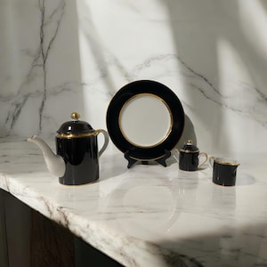 May include: Black and gold tea set with a teapot, creamer, sugar bowl, and a plate. The set is on a white marble surface.