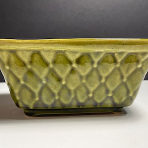 Puede incluir: Un macetero de cerámica verde con un patrón de diamantes. El macetero tiene una forma cuadrada con esquinas redondeadas y una base ligeramente elevada.
