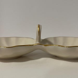 May include: A white porcelain double dish with a gold trim and a handle. The dish is shaped like two scallop shells.