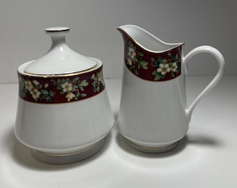 May include: A white porcelain creamer and sugar bowl set with a red and green floral design. The set has a gold trim around the edges.