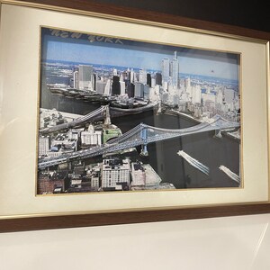 May include: Framed photograph of an aerial view of New York City, featuring the Brooklyn Bridge and the World Trade Center towers. The image is in colour and has a vintage feel.