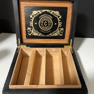 May include: A black wooden cigar box with a gold-colored label that reads "Robusto Larga Camacho C Negro Handmade in Honduras". The box has a hinged lid and a compartment with dividers inside.