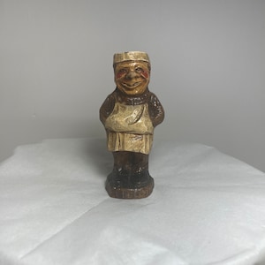 Può includere: Una piccola statuetta dipinta a mano di un uomo che indossa un grembiule bianco e un cappello bianco. La statuetta è in legno e ha una finitura marrone. L'uomo ha un viso sorridente con le guance rosse.