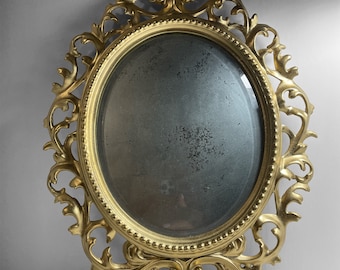 May include: An oval vanity mirror in gold, featuring an elaborate, swirling frame. The mirror has a decorative top and a stand. The mirror's surface shows signs of age, enhancing its vintage look. The frame is made of metal.