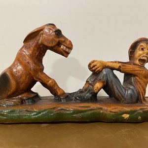 Può includere: Una statuetta di legno di un uomo seduto su un asino. L'uomo indossa un cappello e ha un'espressione sorpresa sul viso. L'asino è marrone e ha una coda lunga.