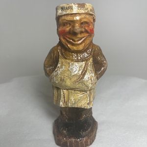 Può includere: Una piccola statuetta dipinta a mano di un uomo sorridente che indossa un cappello bianco con la scritta "Miami Beach, FL". La statuetta indossa una giacca marrone e un grembiule bianco. La statuetta è in piedi su una base marrone.