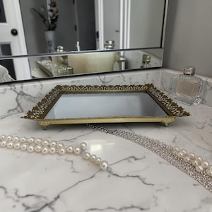 May include: A rectangular, gold-toned vanity tray with a mirrored surface and ornate filigree edges. The tray is resting on a marble countertop, with a perfume bottle and a pearl necklace nearby. The tray is approximately 10 inches long.