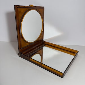 May include: A square, amber-colored plastic folding mirror with a hinged design. The top section features a circular mirror set within a square frame, while the bottom section contains a square mirror. The translucent material has a warm, brownish hue.