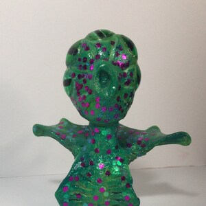 Könnte beinhalten: Eine glitzernde Figur in Grün und Pink mit einem fantasievollen Design. Die Figur hat einen großen Kopf mit einem wirbelnden Muster und einen kleineren Körper mit einem breiten, offenen Kragen.