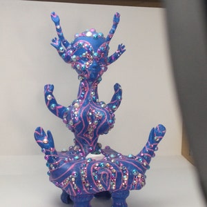 May include: A blue and pink ceramic sculpture of a stylized figure with multiple arms and legs. The figure is covered in rhinestones and has a single eye.