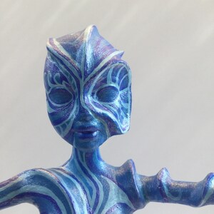 Puede incluir: Una figurita azul y morada con un patrón detallado y en espiral. La figurita tiene una forma humanoide con una cara estilizada y los brazos extendidos.