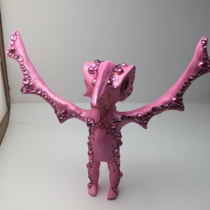 May include: Pink figurine with wings, decorated with rhinestones and gold paint. The figurine has a bird-like head and is standing with its wings outstretched.