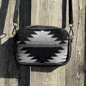 May include: Black and gray patterned crossbody bag with a black leather trim and strap. The bag features a geometric design in shades of black and gray. The bag has a gold zipper and metal hardware.