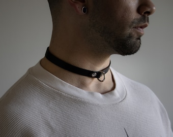 May include: Black leather choker necklace with a silver-colored ring and hardware. The choker is worn around the neck. The background is a plain, light gray wall. The person is wearing a white waffle-knit shirt.