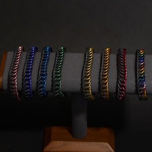 May include: A display of colorful chainmail bracelets. The bracelets are arranged on a gray stand, showcasing a variety of colors including red, blue, green, and gold. The bracelets are made of small, interlocking metal rings.