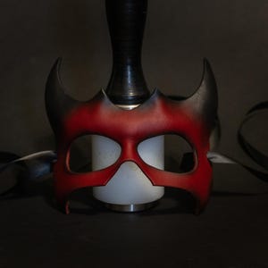 May include: A red and black leather mask with a horned design. The mask has eye cutouts and is resting on a white cylindrical object. A black strap is draped around the mask.