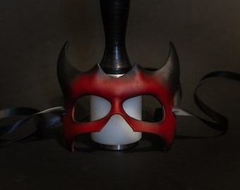 May include: A red and black leather mask with a horned design. The mask has eye cutouts and is resting on a white cylindrical object. A black strap is draped around the mask.